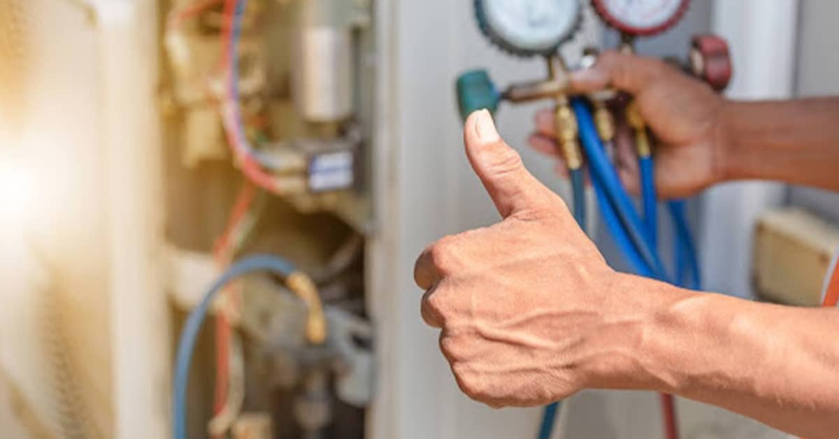 A technician gives a thumbs up with his left hand while working on an HVAC system. He holds a blue and red manifold gauge in his right hand.