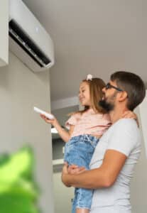 Happy little girl holds remote control for ductless air conditioner sitting in fathers hands in light interior of apartment.