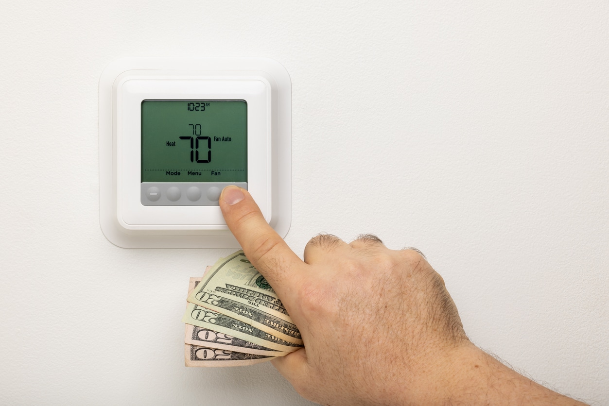 A homeowner’s hand holds 20 dollar bills as he adjusts a thermostat on a wall for more energy-efficient heating.
