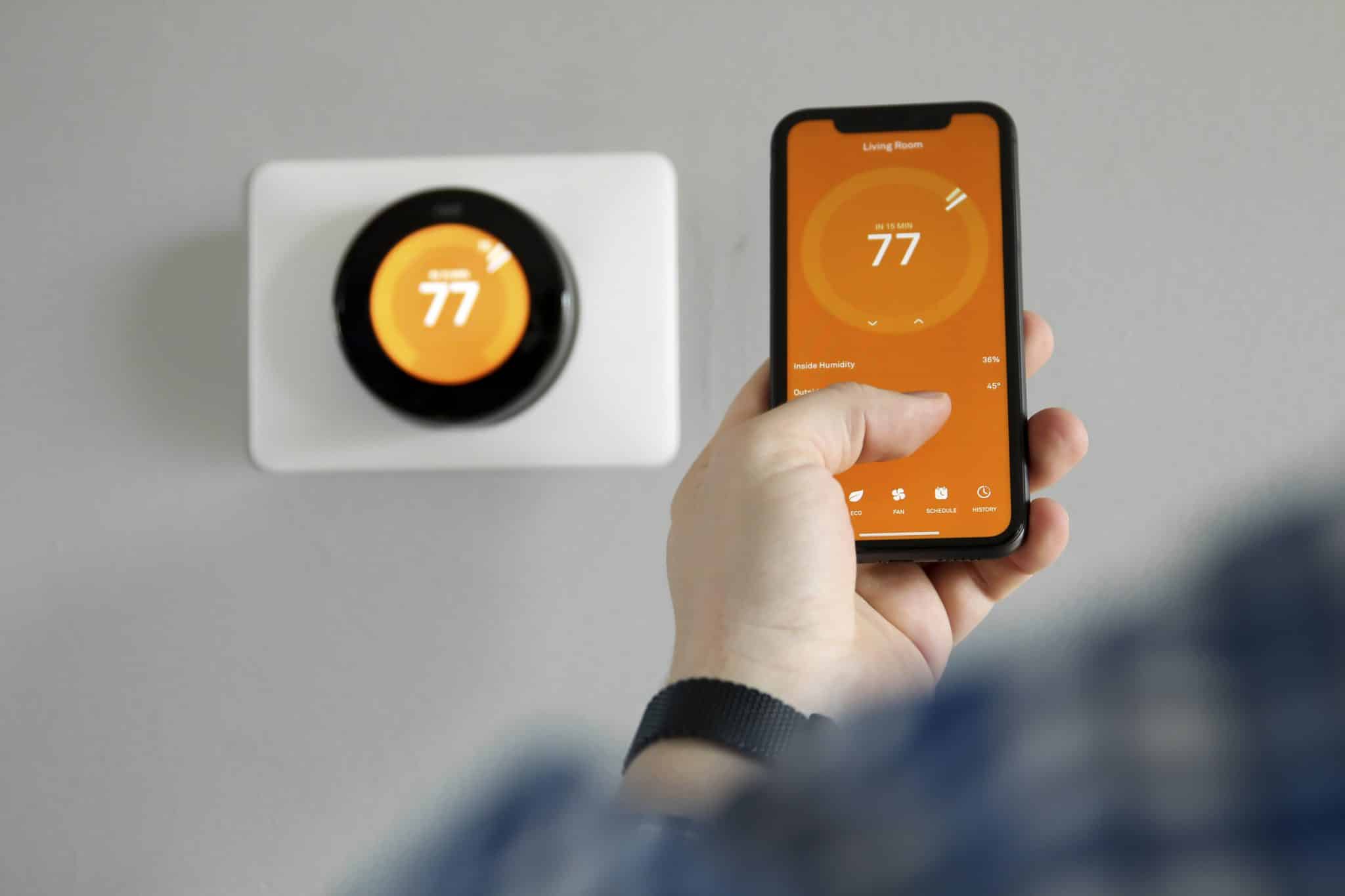 A smart home thermostat on the wall, reading 77, and someone holding a phone with a similar temperature display.