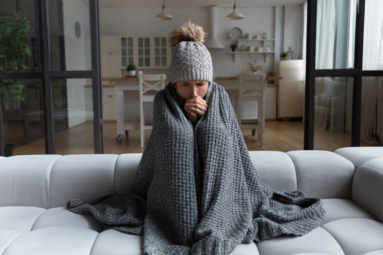Person bundled in a gray blanket and knit hat on couch, feeling cold at home after skipping her heater maintenance plan.