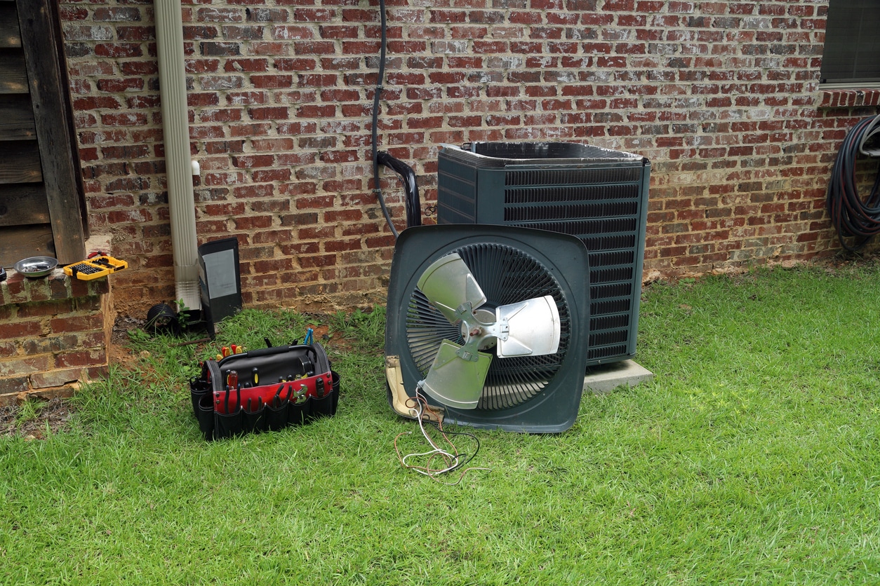Air conditioning unit repair in progress with tools on grass beside a brick wall.